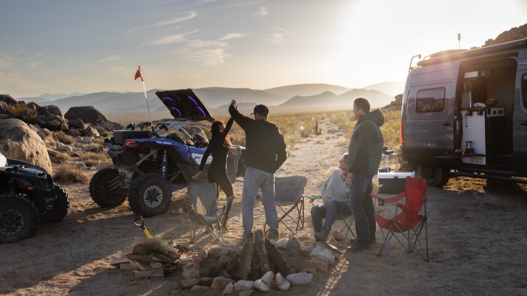 A group of four people on camping chairs by a fire pit dancing, wiith a Can-Am Side-by-Side and Van, plus mountains in the background