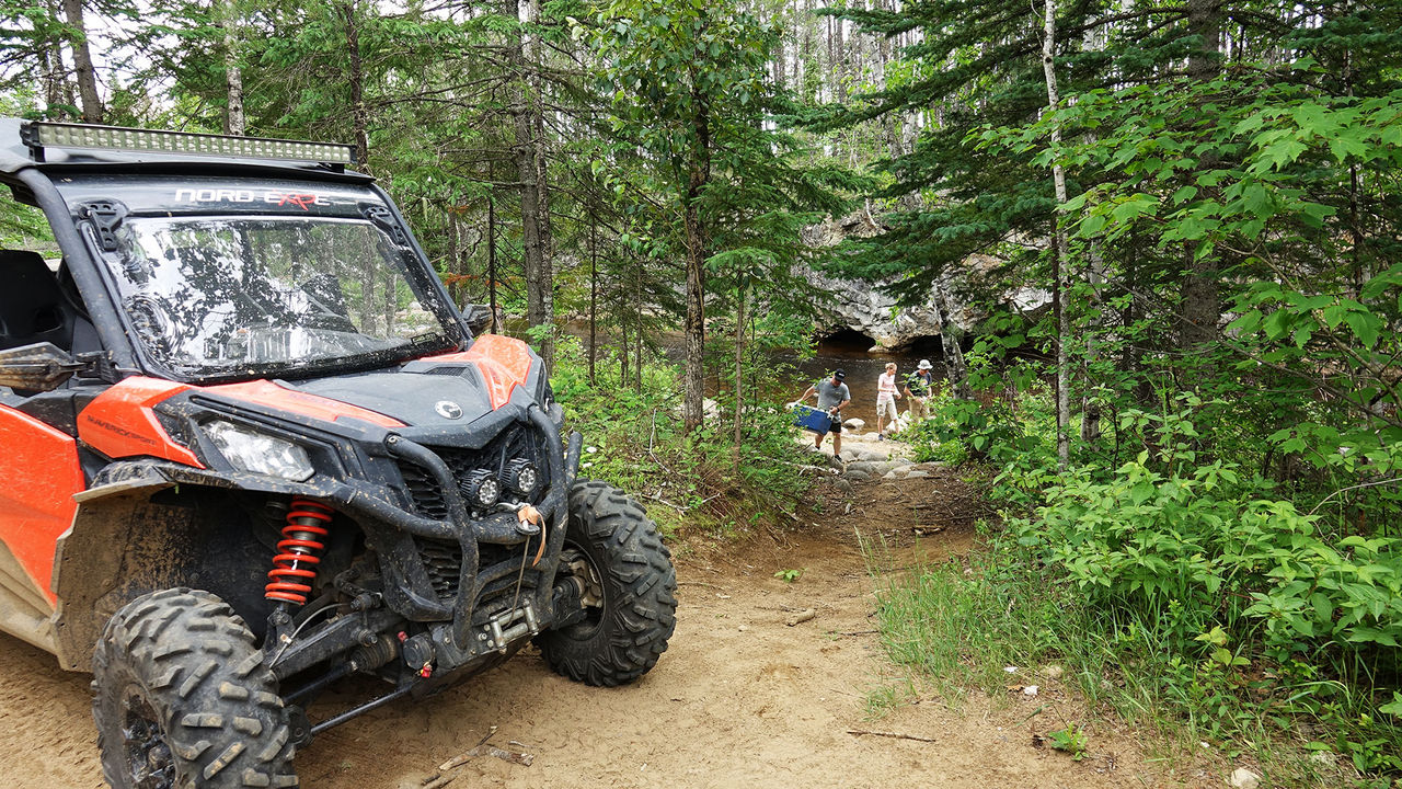 Can-Am Side-by-Side parked, with 3 people in the background (one carrying a cooler), behind some trees, near flowing water. 
